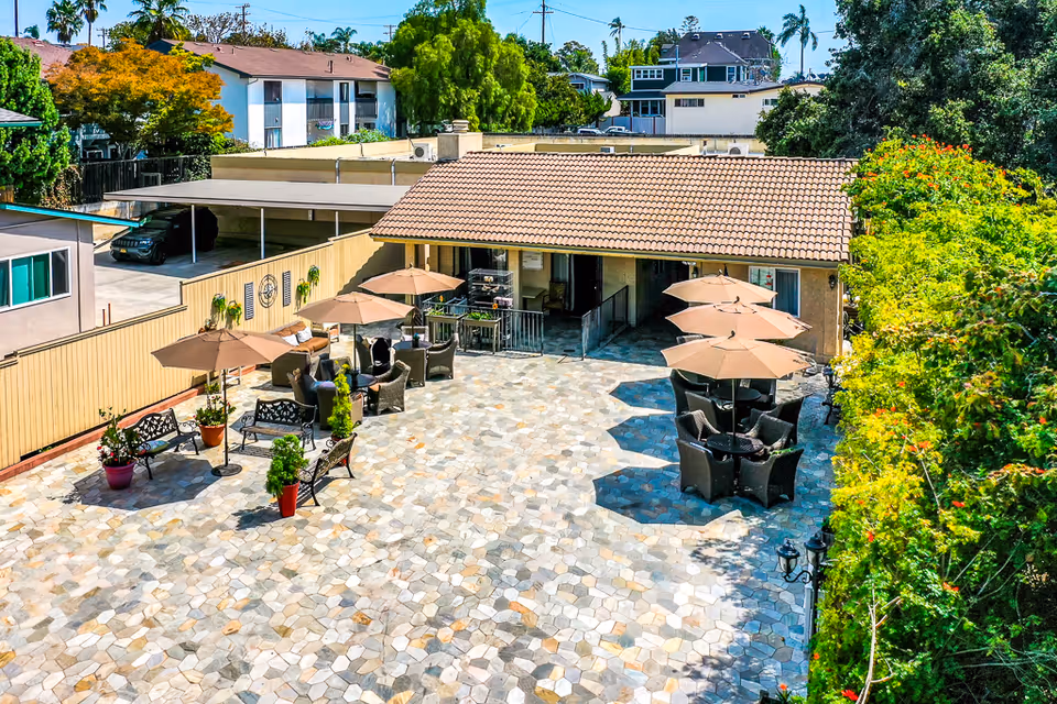 Outdoor patio area with several tables and chairs under large beige umbrellas, surrounded by greenery and adjacent to a building with a tiled roof. There are benches and potted plants along a beige fence, and a covered parking area with cars is visible in the background.