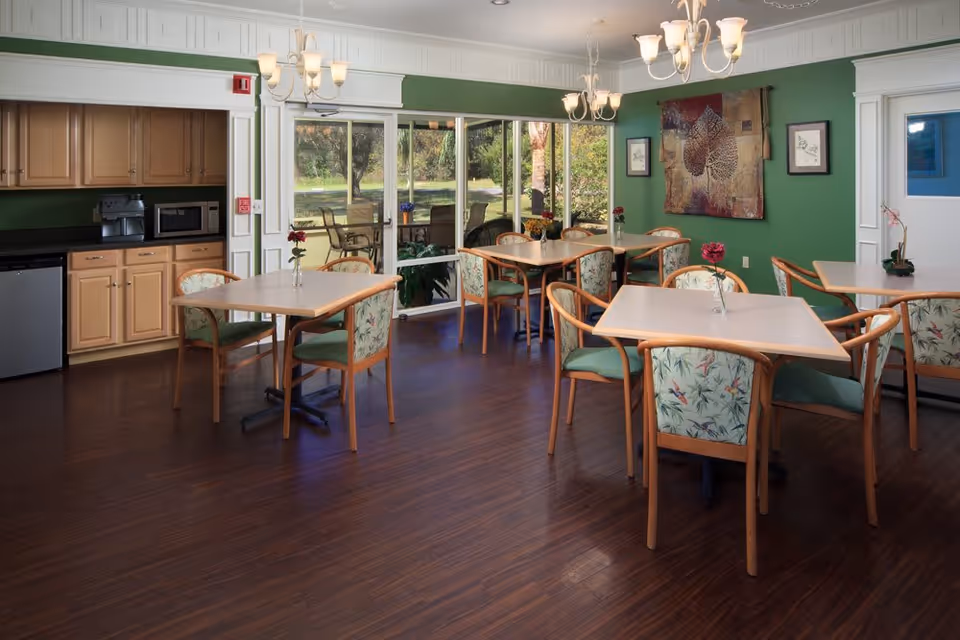 A dining room with several square tables and wooden chairs with floral upholstery. The room has green walls, dark wood flooring, and chandeliers hanging from the ceiling. There is a kitchenette area with cabinets, a microwave, and a coffee machine on the left side. Large sliding glass doors lead to an outdoor patio with additional seating and greenery visible outside.