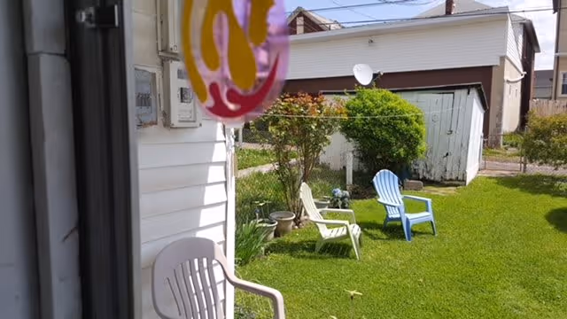 View of a small backyard with green grass, two plastic lawn chairs (one white and one blue), a small tree, and a white shed with a brown roof in the background. Part of a white house and a window frame are visible on the left side of the image.