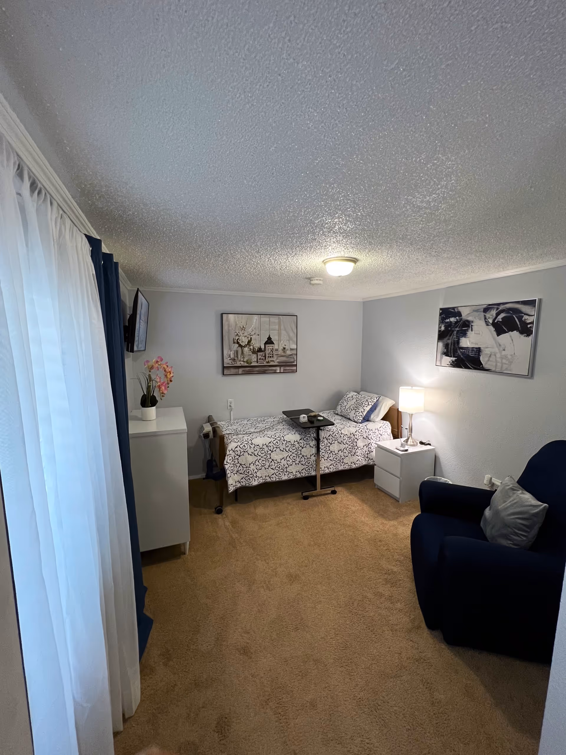 Small furnished bedroom with a single bed, bedside table and lamp, armchair, dresser, and wall art.