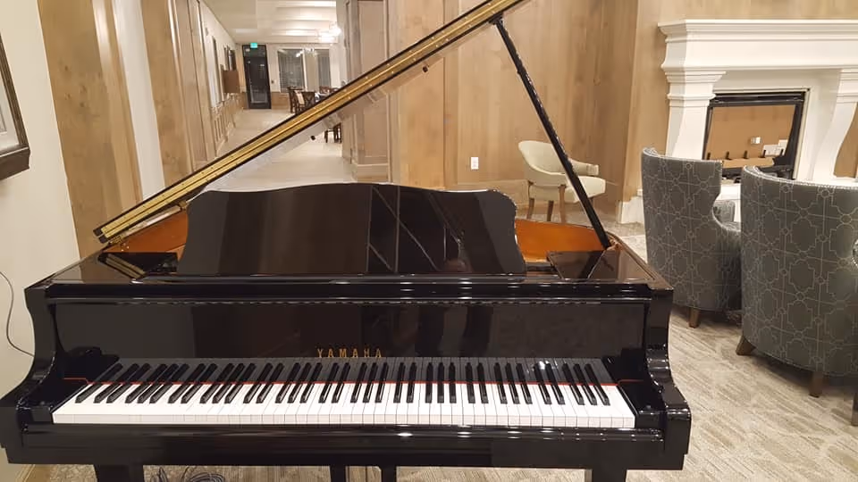 A black Yamaha grand piano in the foreground of a well-lit room with beige walls and carpet. Behind the piano, there are two patterned armchairs facing a white fireplace. A hallway with additional seating is visible in the background.