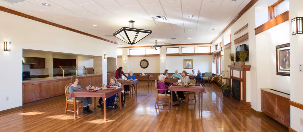 Bright dining room with wooden floors, tables where residents are seated, an open serving counter, and a fireplace.