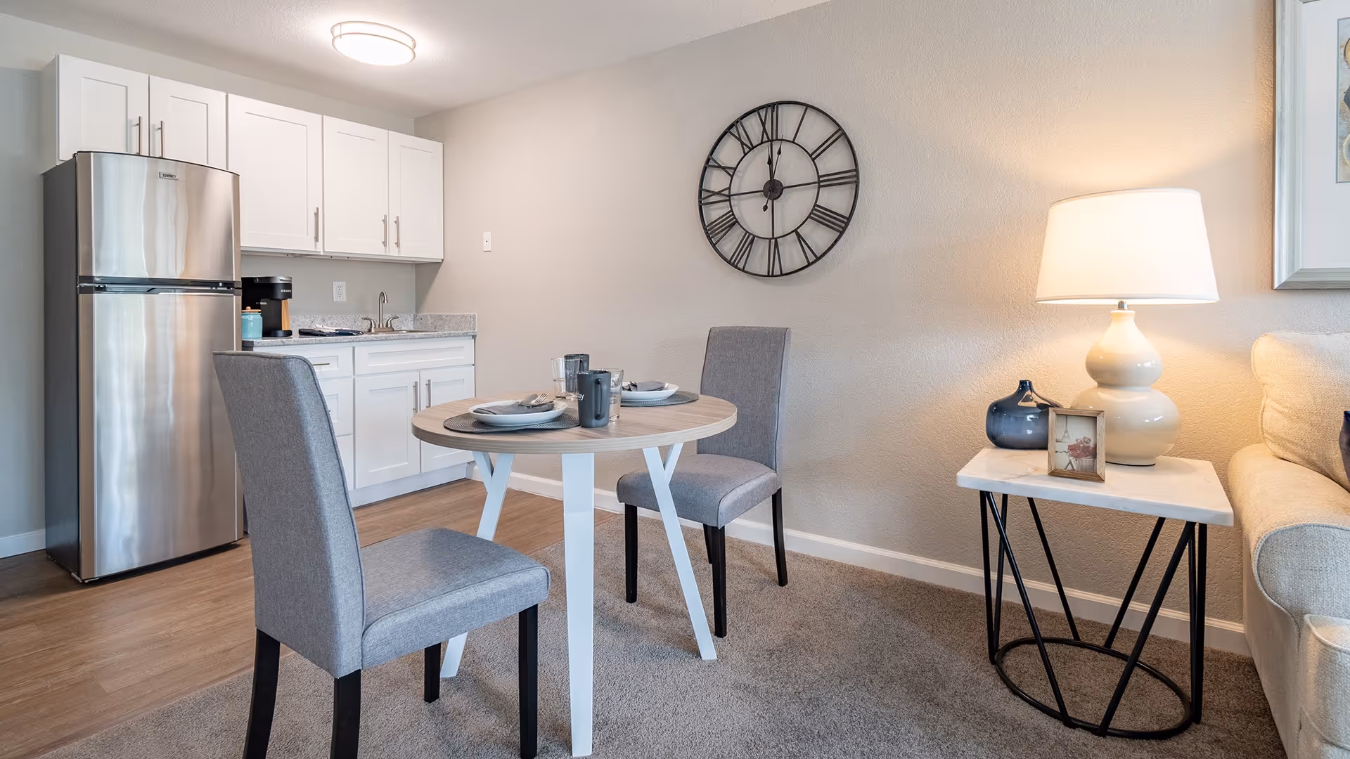 A small dining area in a retirement community apartment with a round wooden table set for two, two gray upholstered chairs, a white side table with a lamp and decorative items, a beige couch, and a kitchenette with white cabinets and a stainless steel refrigerator in the background.