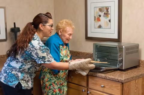 A caregiver helps an elderly woman remove a baking tray from a countertop toaster oven in a small kitchen area.