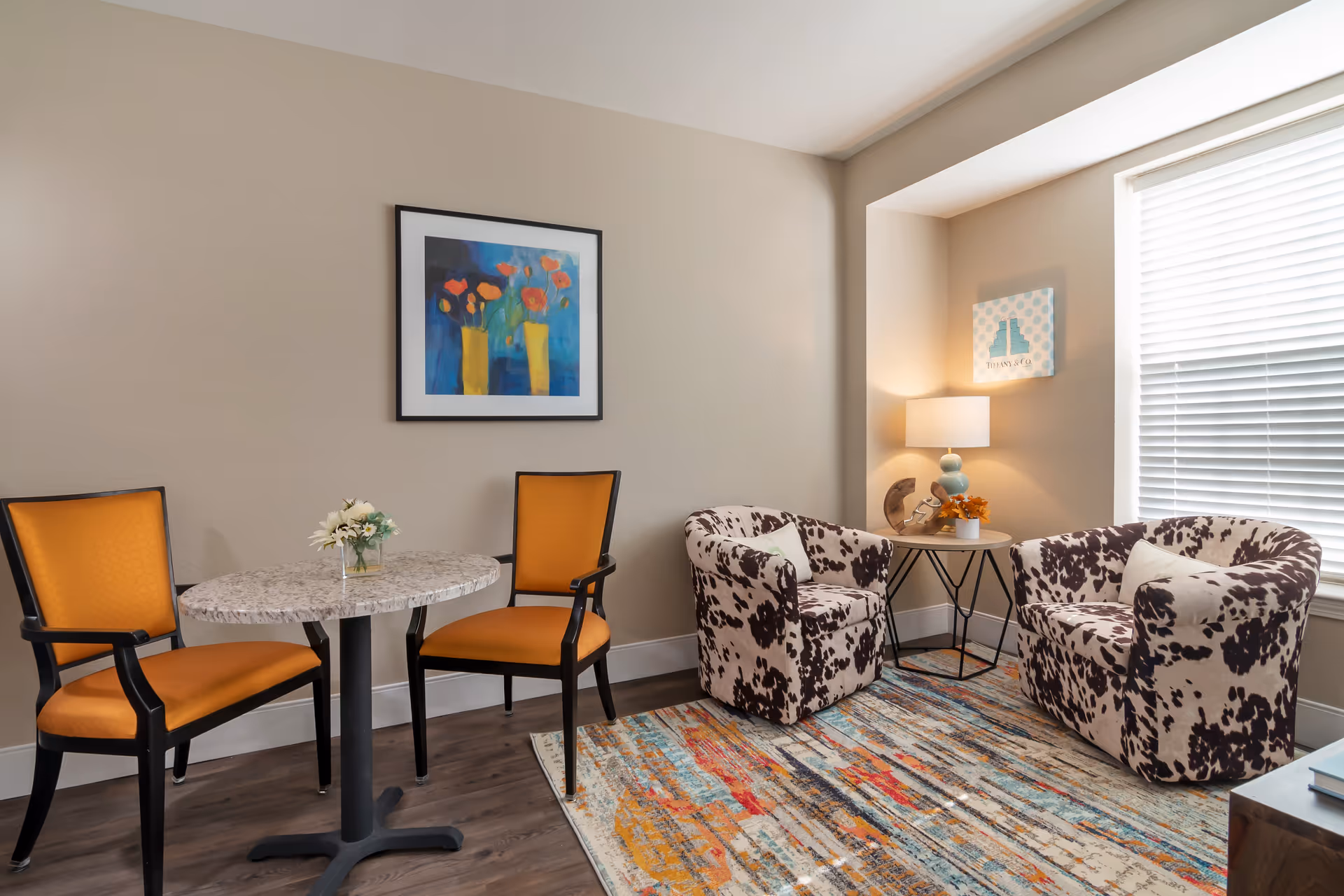A cozy sitting area with two orange cushioned chairs around a small round table with a flower vase, and two patterned armchairs with a small side table and lamp between them, next to a window with blinds. A colorful abstract painting hangs on the wall above the orange chairs.