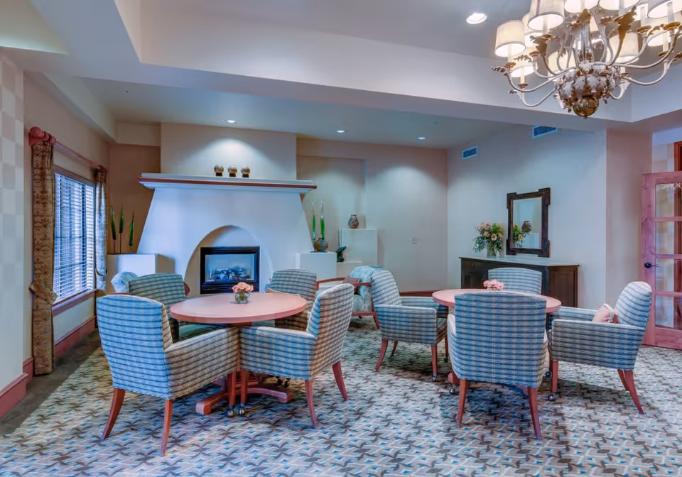 A cozy interior room with two round tables, each surrounded by four upholstered chairs with a blue and white pattern. The room features a decorative fireplace in the background, a window with blinds and curtains on the left, a wooden sideboard with a mirror and flower arrangement on the right, and a chandelier hanging from the ceiling.