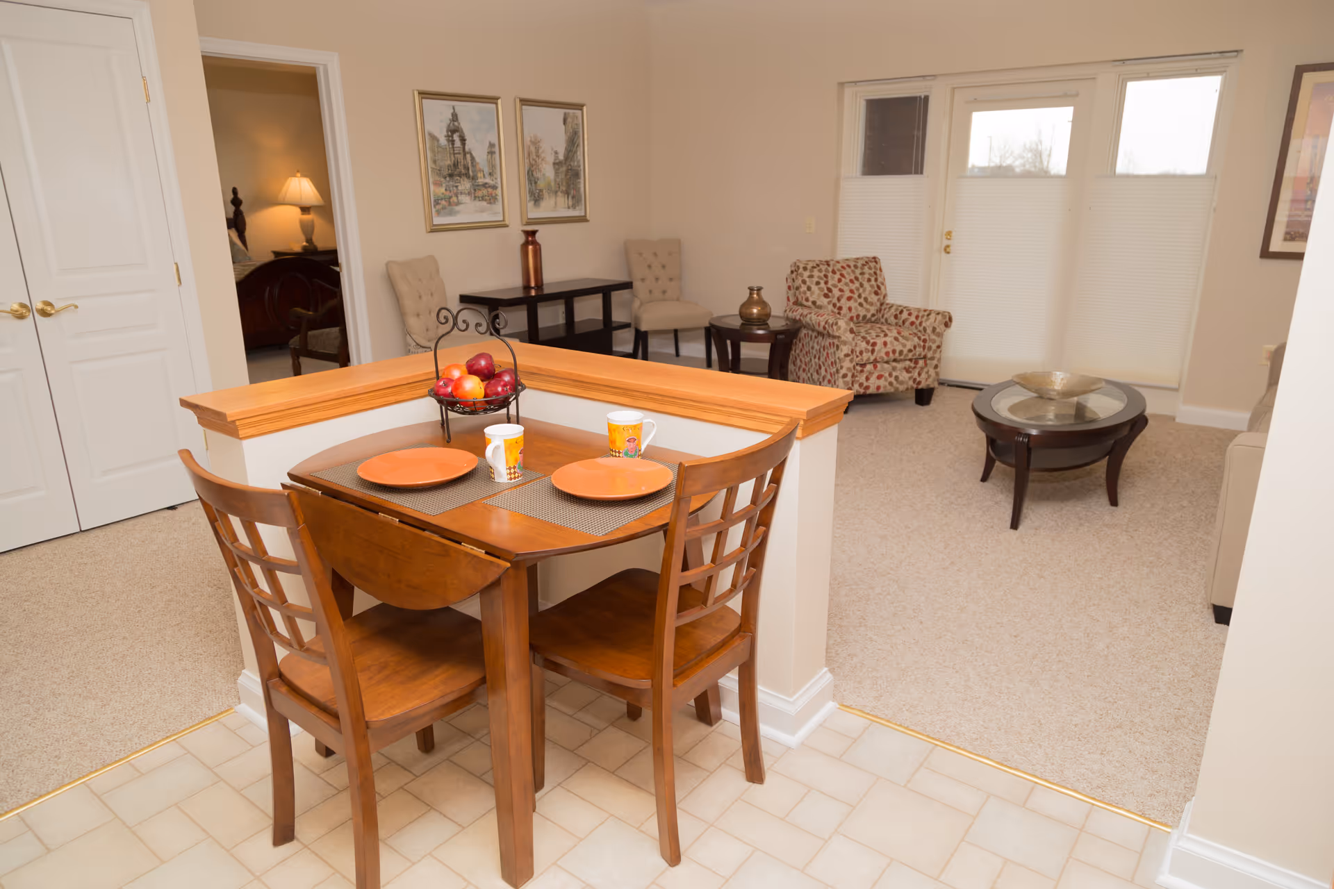 A cozy interior space featuring a small wooden dining table set for two with orange plates and mugs, a fruit basket with apples on the table, and a living area with beige carpet, a patterned armchair, a beige sofa, and a glass-top coffee table. There are two framed pictures on the wall and a door leading to a bedroom with a lit lamp visible.