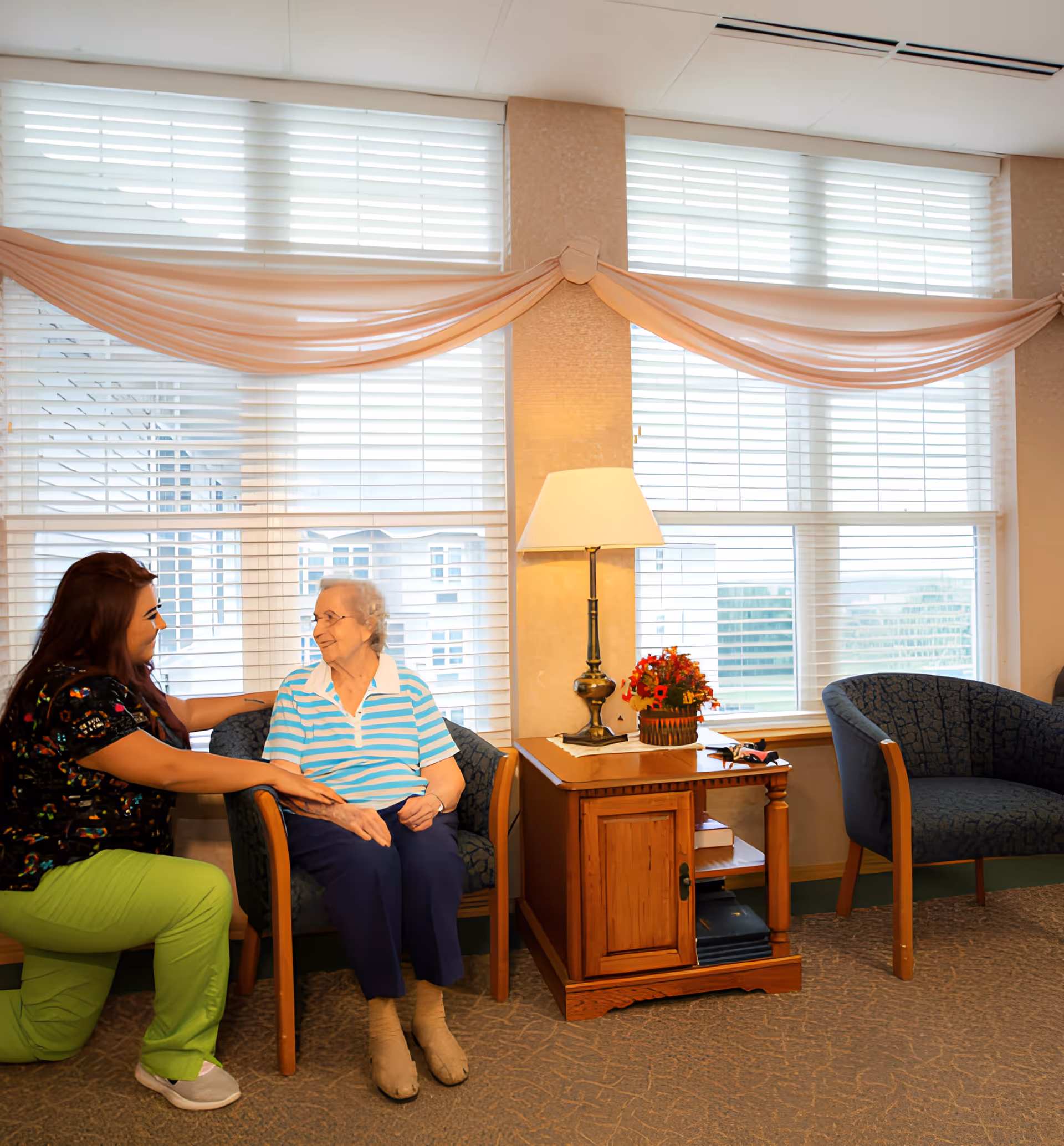 A caregiver kneeling beside an elderly woman seated in a chair in a well-lit room with large windows covered by white blinds and peach-colored drapes. There is a wooden side table with a lamp and a small flower arrangement between two blue upholstered chairs.