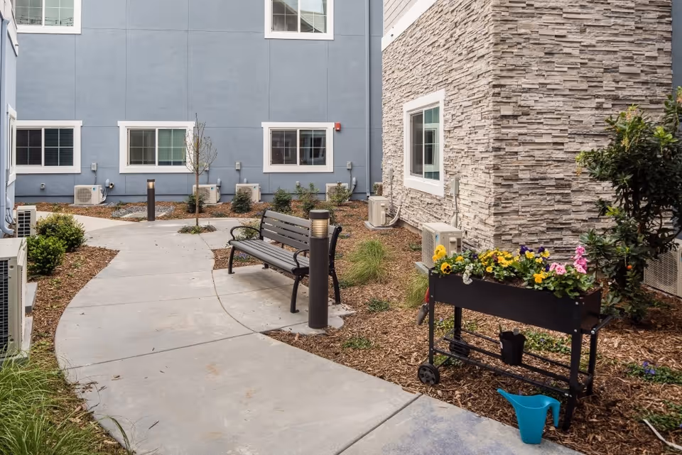Small courtyard with a curved concrete path, a bench, a wheeled planter of flowers, and surrounding apartment building walls with windows.