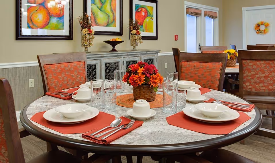 A round dining table set for six with white plates, bowls, cups, silverware on red napkins, and clear drinking glasses. A basket with red and orange flowers is in the center. The room has wooden chairs with orange patterned cushions, colorful fruit paintings on the wall, and a sideboard with decorative items.