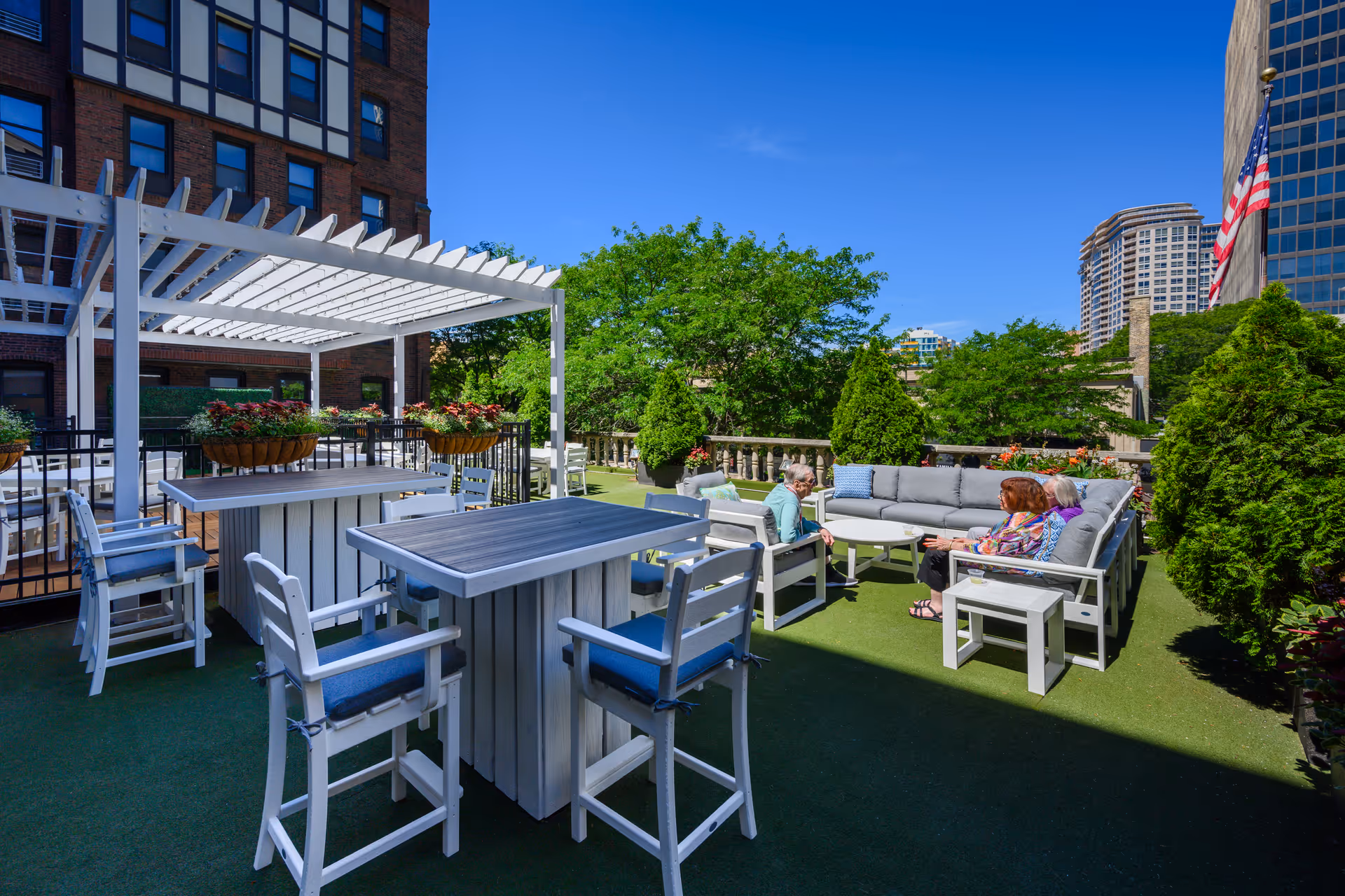 Outdoor patio area at The Merion with white wooden tables and chairs, a white pergola, potted plants, and a group of elderly people sitting on gray cushioned outdoor sofas under a clear blue sky with surrounding greenery and city buildings in the background.