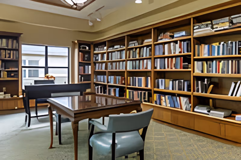 A quiet library room with wooden bookshelves filled with books and binders lining the walls. There is a wooden table with a glass top in the center, surrounded by chairs. A window lets in natural light, and the room has a calm, studious atmosphere.