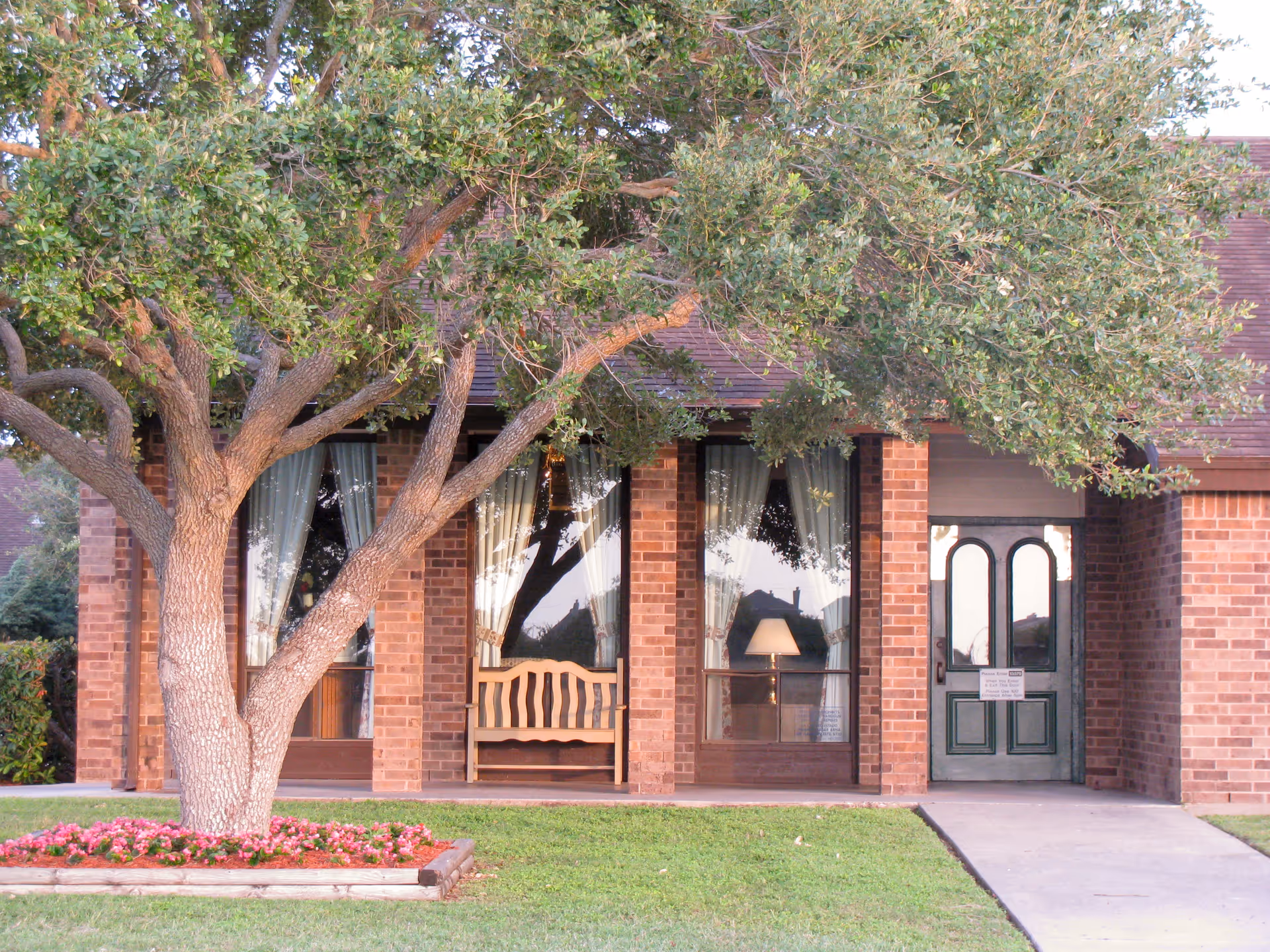 Front entrance of a brick nursing center with a large tree, a bench between tall windows, and a green double door.