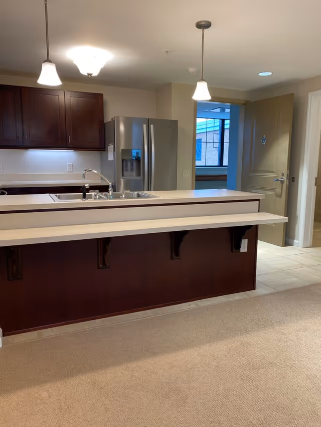Interior view of a kitchen area in a senior living facility featuring a double sink with a chrome faucet on a beige countertop. Above the sink are two pendant lights and a ceiling light. Behind the sink is a stainless steel refrigerator and dark wooden cabinets. To the right, there is an open door leading to another room with a window visible.