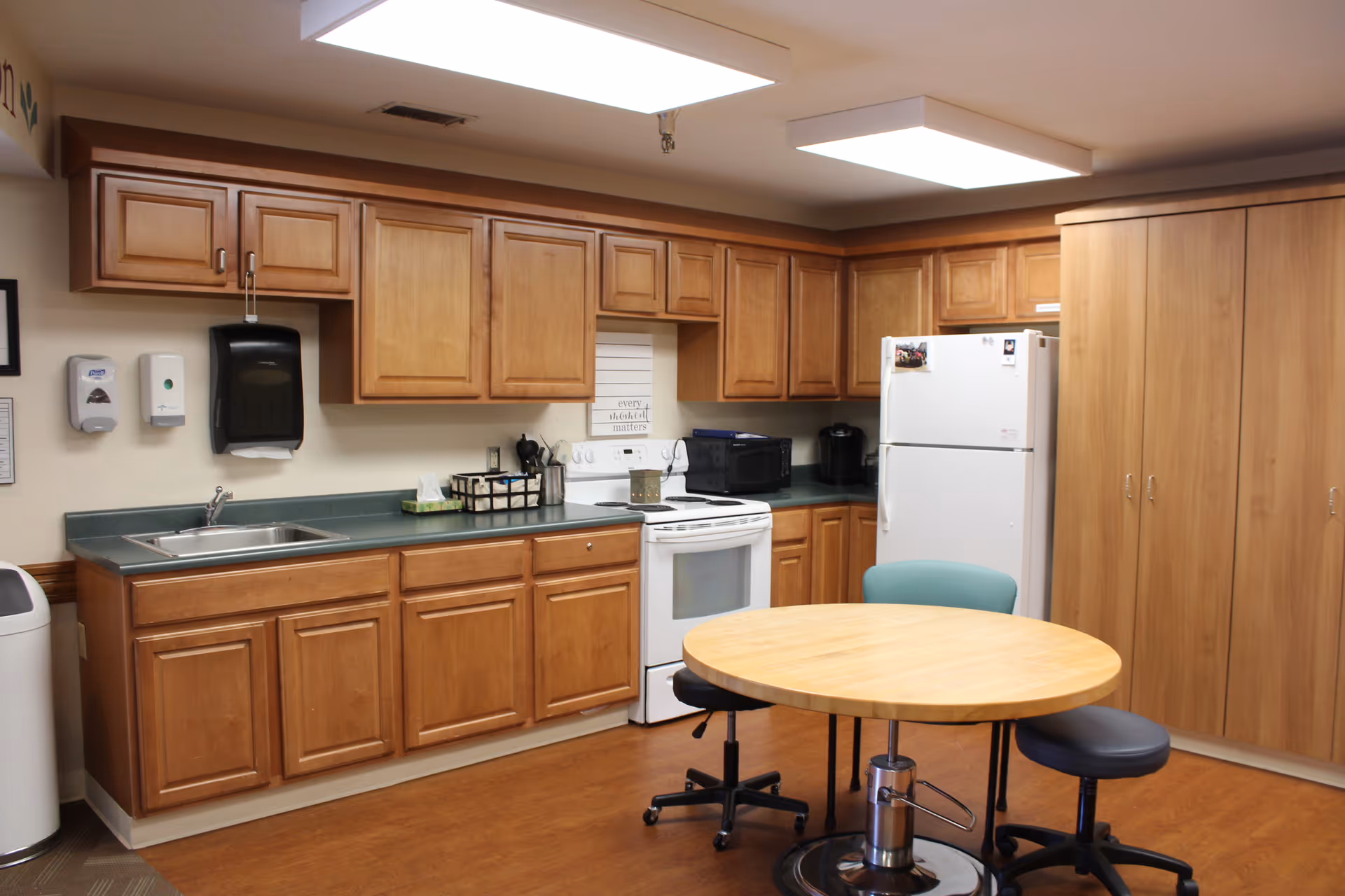 A kitchen area with wooden cabinets, a white stove, a white refrigerator, a microwave, a coffee maker, a sink, and a round wooden table with two chairs on wheels. The room has overhead fluorescent lighting and a wood floor.