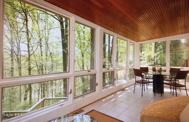 A bright enclosed porch or sunroom with large windows overlooking a wooded outdoor area. The room features a tiled floor, a round glass table with four chairs, and a wicker chair in the foreground. The ceiling is wooden with a warm finish.