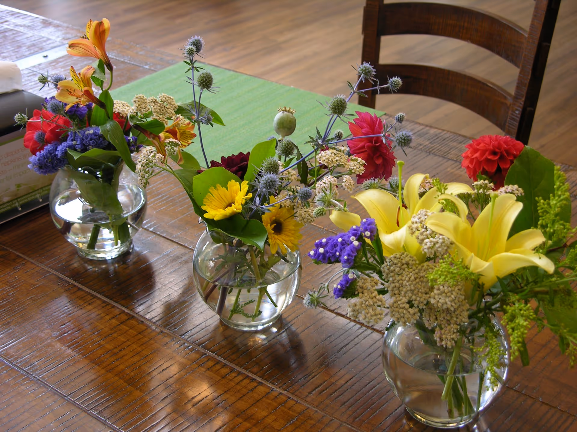 Three small glass vases with colorful flower arrangements placed on a wooden table with a green table runner, with a wooden chair and wooden floor visible in the background.