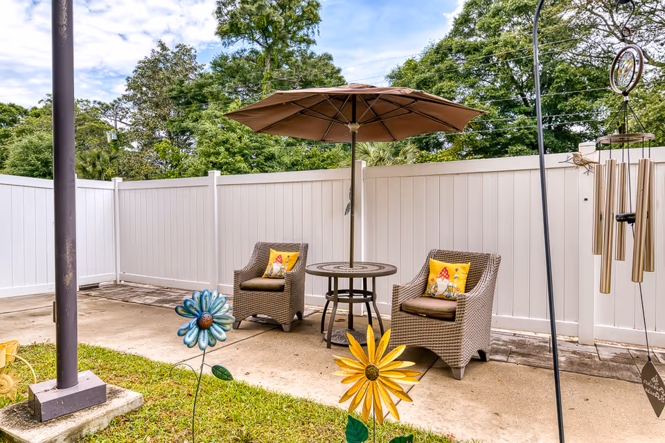 Outdoor patio area with two wicker chairs each with a yellow pillow featuring a gnome design, a round table with a large brown umbrella, white privacy fence in the background, decorative metal flowers in the foreground, and a wind chime hanging on the right side.