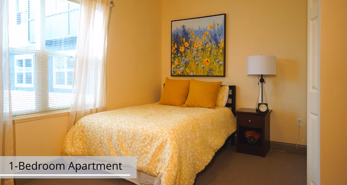 A cozy bedroom with a bed covered in a yellow patterned comforter and two matching pillows. There is a nightstand with a lamp and a clock on it next to the bed. A colorful floral painting hangs on the wall above the bed. Light filters in through a window with sheer curtains.