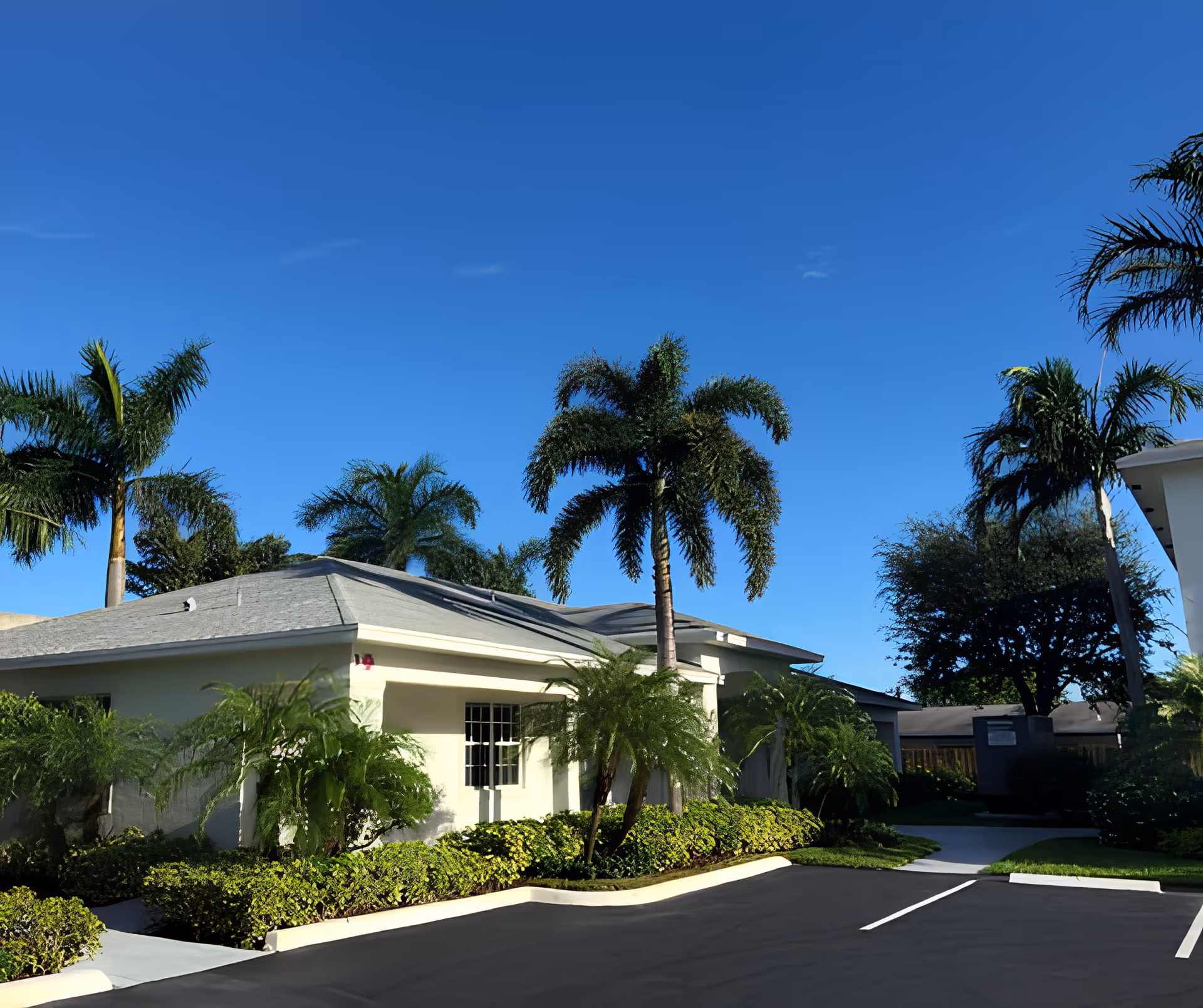 Exterior view of a single-story building with a gray roof surrounded by palm trees and green shrubs under a clear blue sky. There is a paved parking area in the foreground with marked parking spaces.