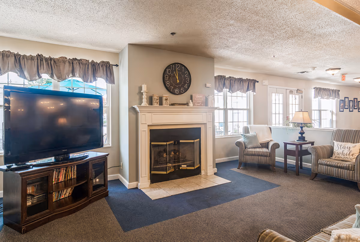 A cozy living room area featuring a large flat-screen TV on a wooden stand with shelves of DVDs, a white fireplace with decorative items and a clock above it, two striped armchairs with pillows, a side table with a lamp, and large windows with curtains letting in natural light.