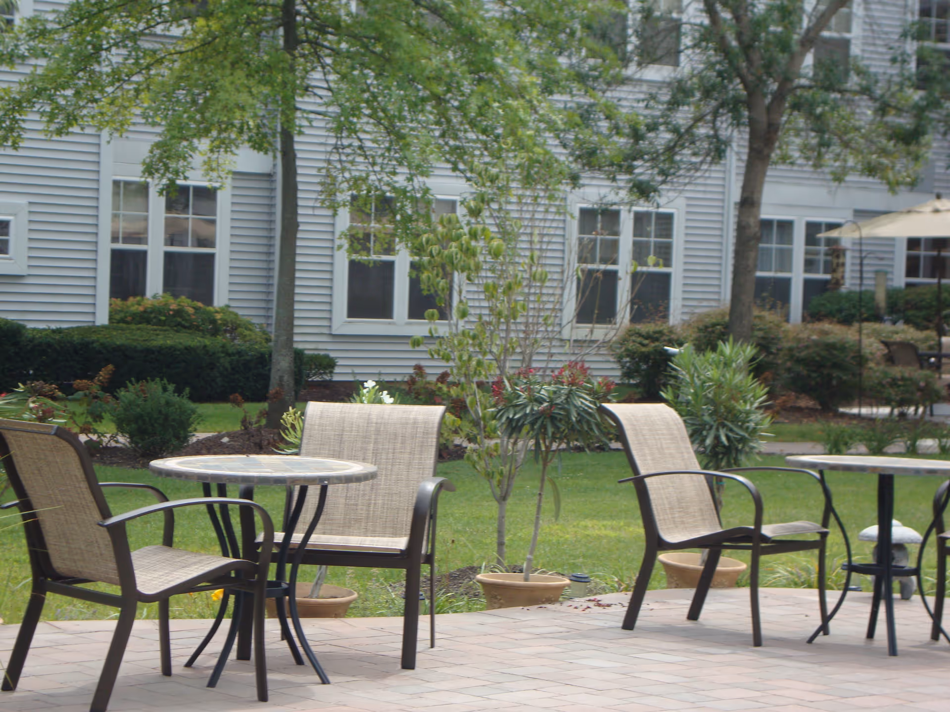 Outdoor patio area with round tables and beige mesh chairs on a paved surface, surrounded by green grass, small trees, and shrubs in front of a light gray building with multiple windows.