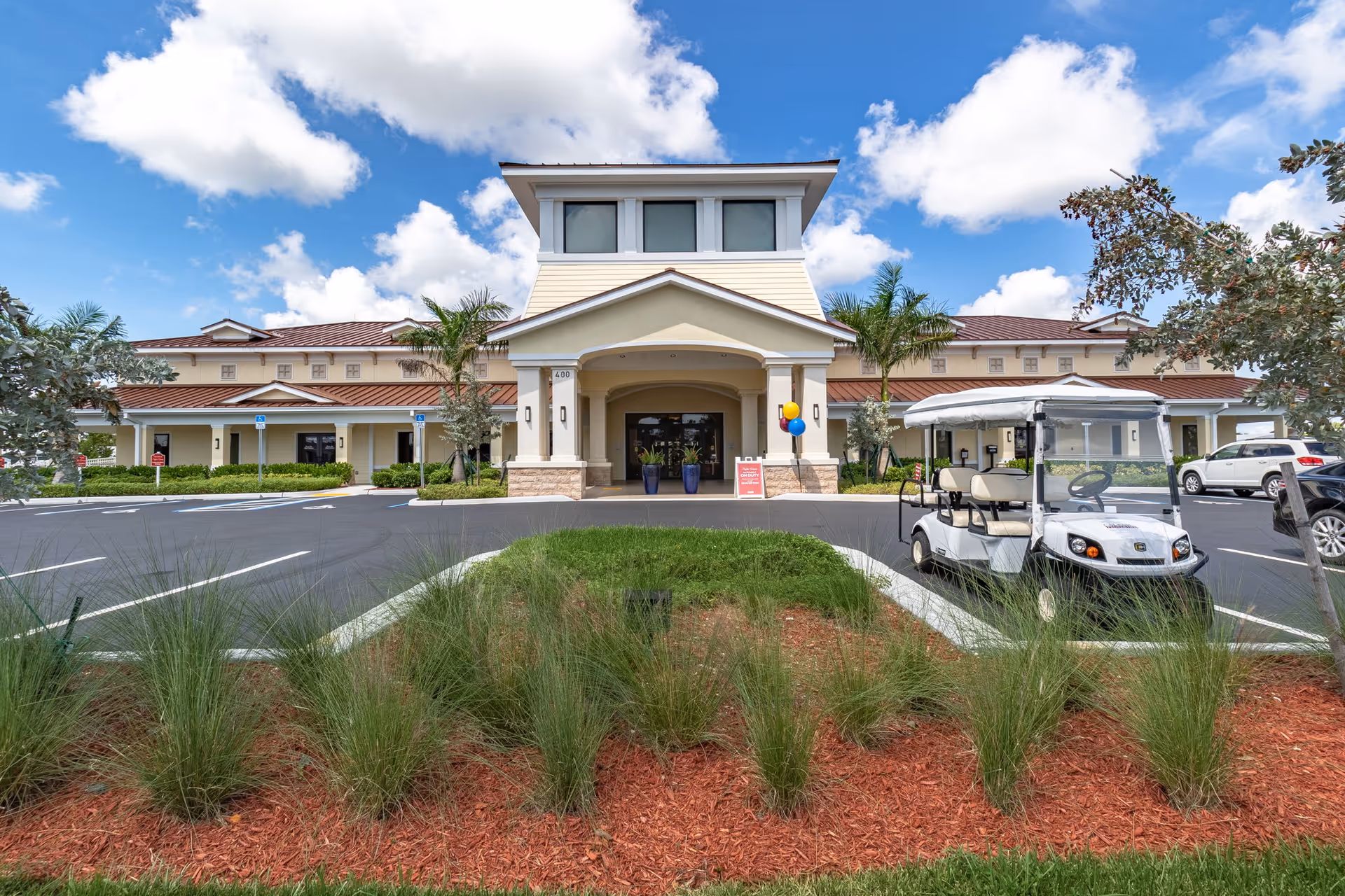 Front exterior view of a single-story building with a covered entrance, palm trees, and a golf cart parked in the parking lot under a partly cloudy blue sky.