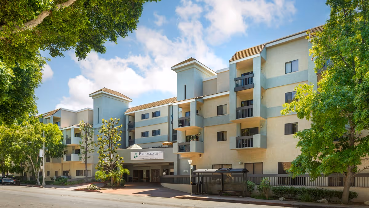 Exterior view of a multi-story senior living facility building with balconies, surrounded by green trees under a partly cloudy sky. The entrance has a sign that reads 'Brookdale Uptown Whittier'.