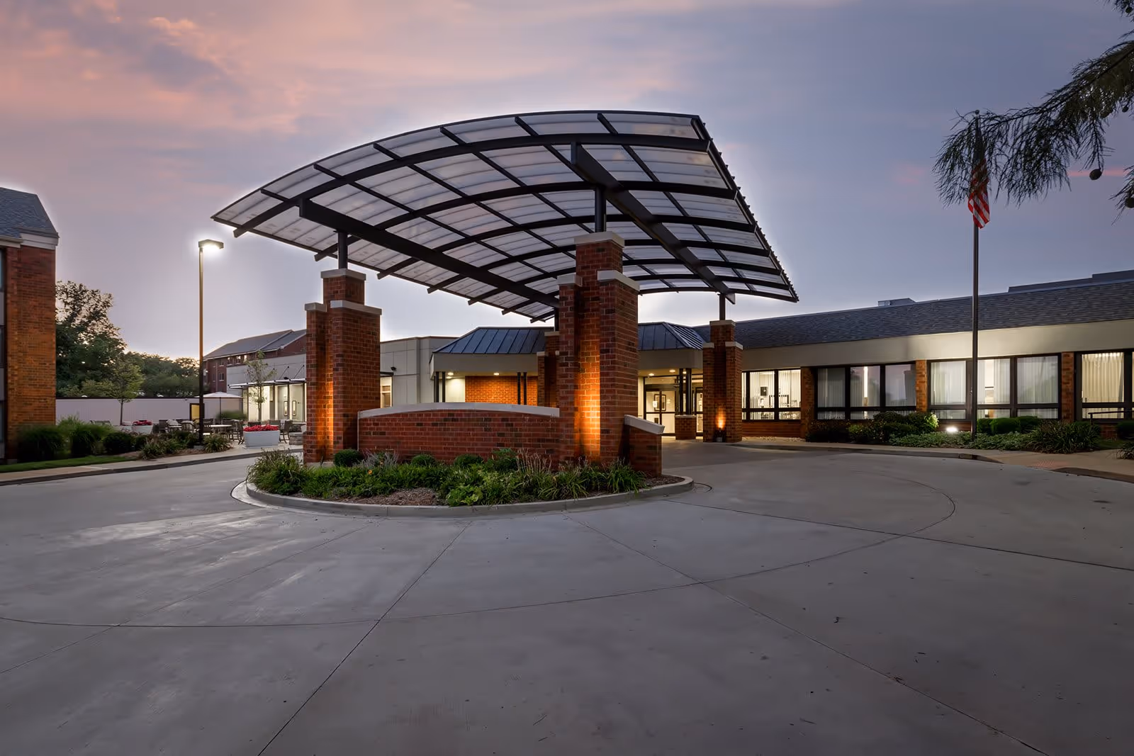 Entrance of Gambrill Gardens Senior Apartments at dusk with a large covered driveway supported by brick pillars, surrounded by well-maintained landscaping and a flagpole with an American flag.