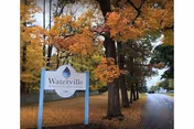 A sign for Waterville Residential Care Center is positioned beside a tree-lined road with autumn foliage. The trees have orange and yellow leaves, and the road extends into the distance on the right side of the image.