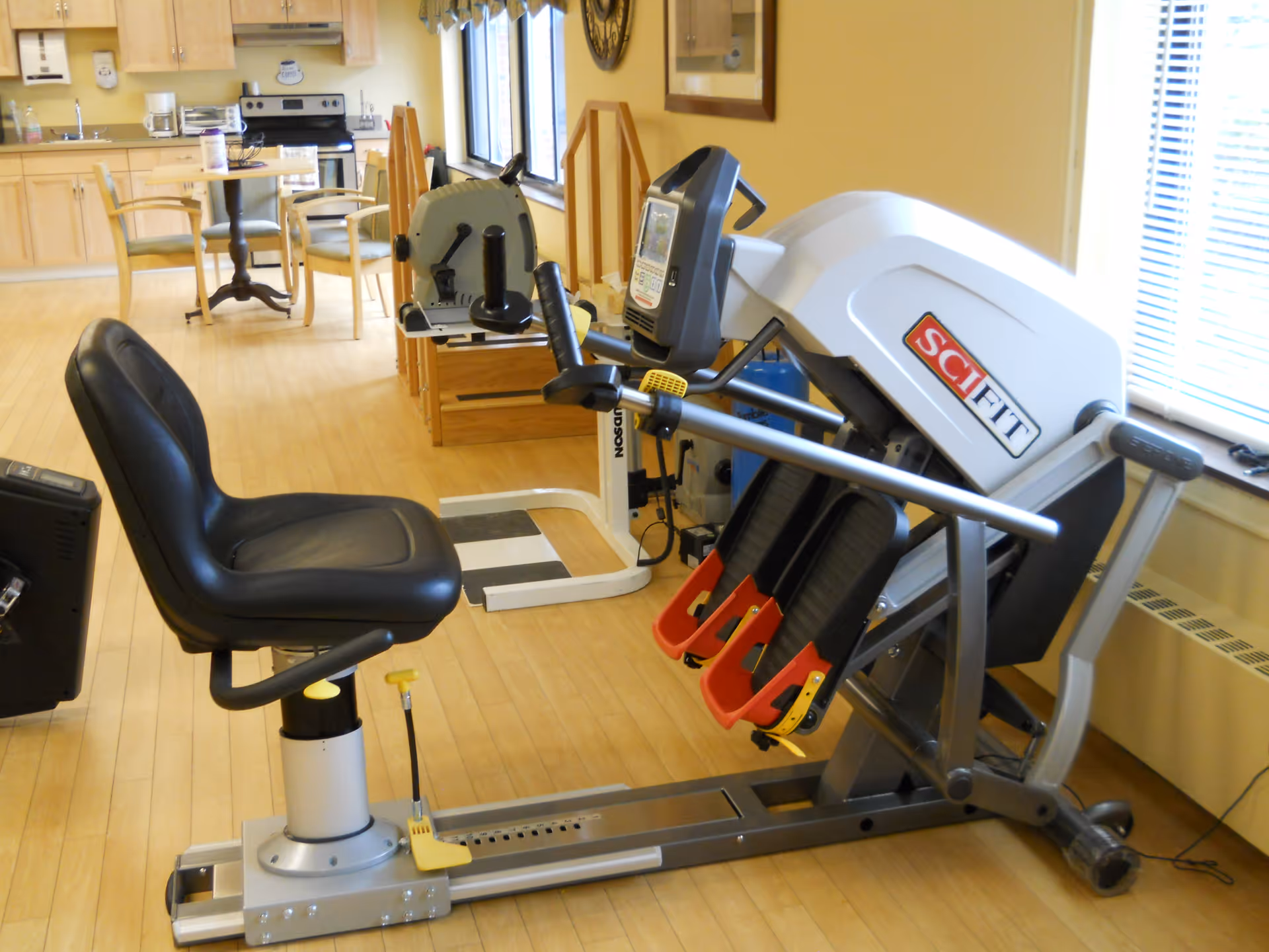 Rehabilitation exercise machines and a recumbent bike in a bright communal room with a kitchenette and table in the background.