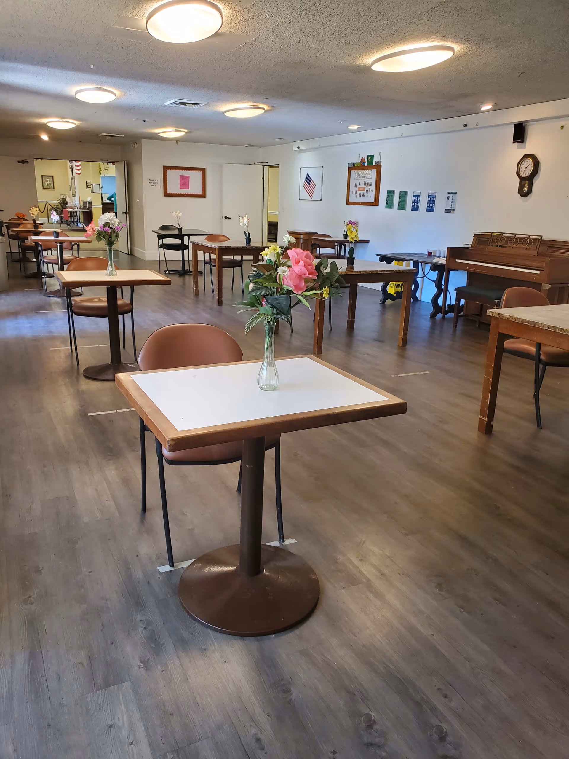 A spacious dining room with several small square tables, each with a single chair and a vase of flowers on top. The room has wooden flooring, white walls decorated with framed pictures and an American flag, and ceiling lights. There is a piano against one wall and a clock hanging above it.