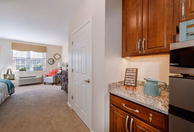 Interior view of a senior living facility room showing a kitchenette area with wooden cabinets and granite countertop on the right, and a living/sleeping area with a bed, armchair, dresser, and window with valance on the left.