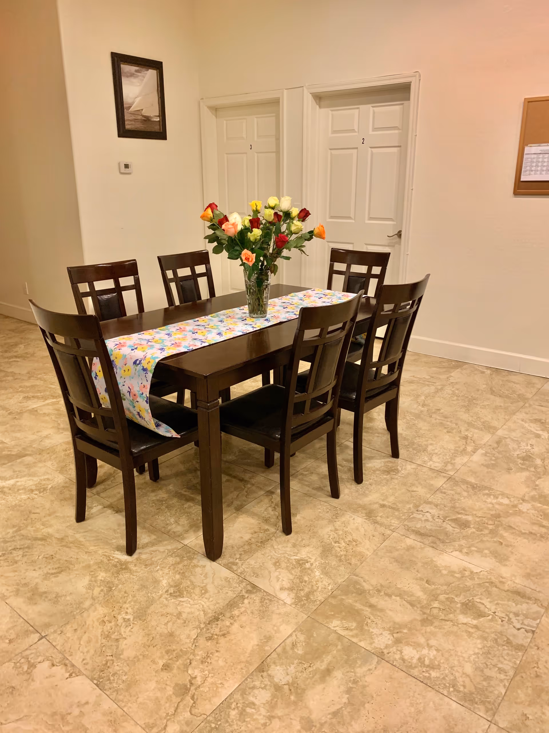 Wood dining table with six chairs and a vase of multicolored roses in a tiled room with numbered doors.