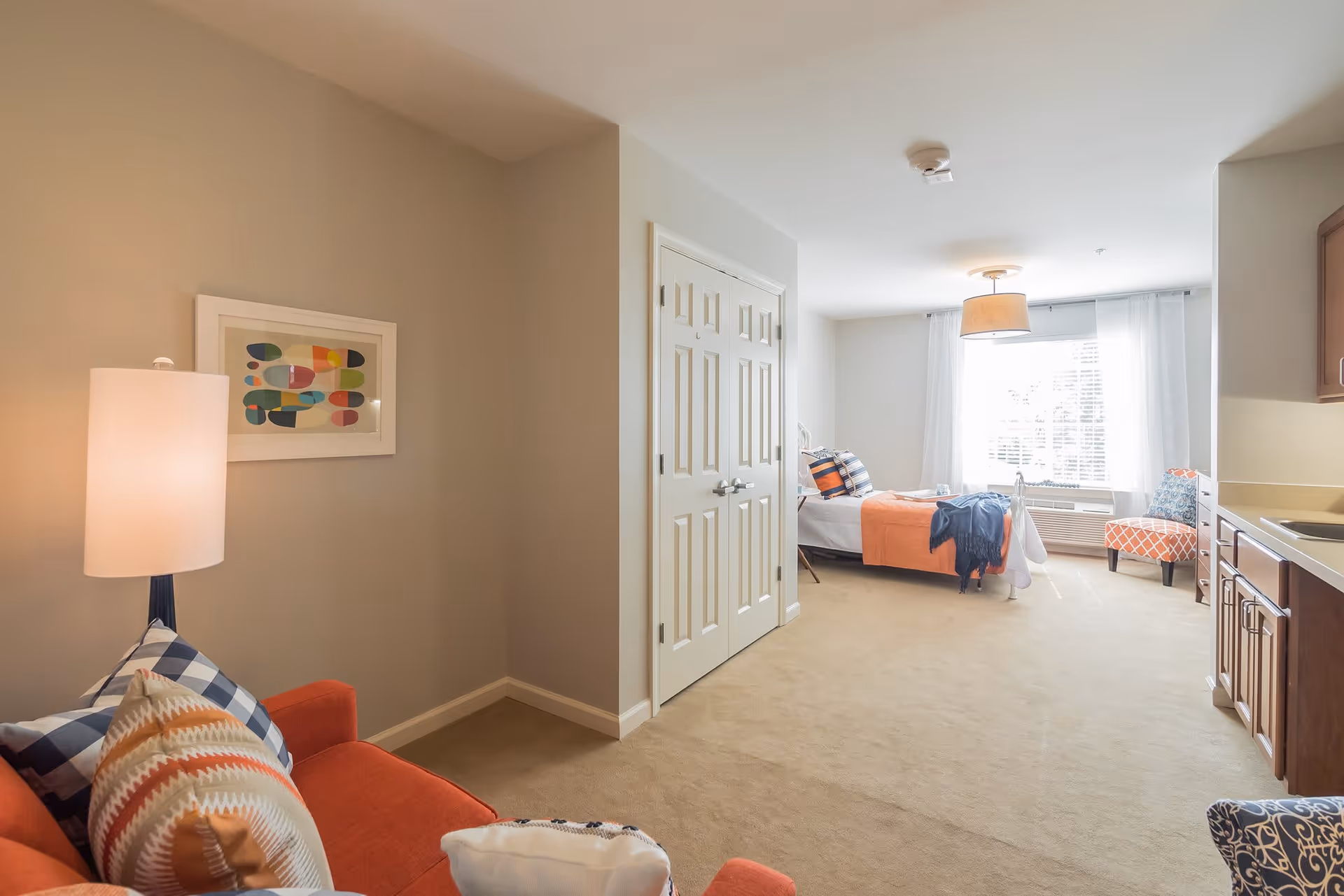 A bright senior living apartment interior featuring a bed with orange and blue bedding near a large window with white curtains. There is a small sitting area with an orange armchair and patterned pillows, a lamp, and a framed abstract artwork on the wall. A kitchenette with wooden cabinets and a sink is visible on the right side.