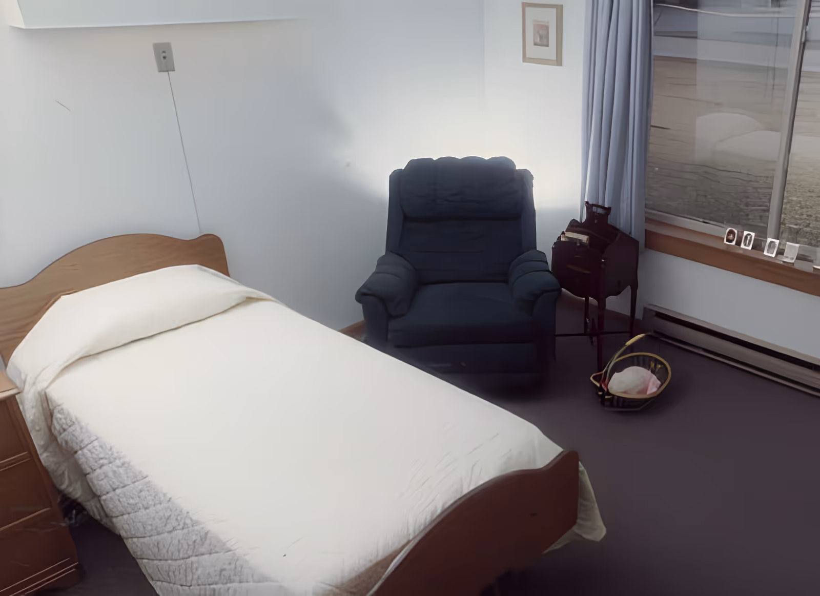 A simple bedroom with a single bed covered in a white quilt, a wooden nightstand, a dark blue armchair, a small wooden side table, and a window with light gray curtains. There is a basket on the floor near the window.