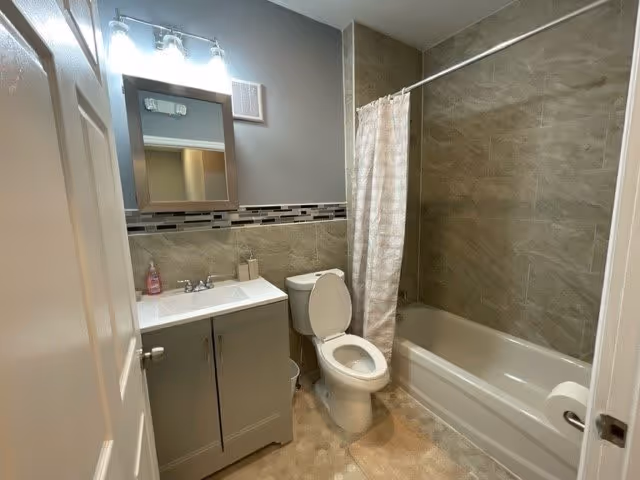 A clean bathroom with a white toilet, a gray vanity with a white sink, a mirror with lights above it, and a bathtub with a beige shower curtain. The walls are tiled in a beige color with a decorative tile border, and the floor has a light-colored pattern.