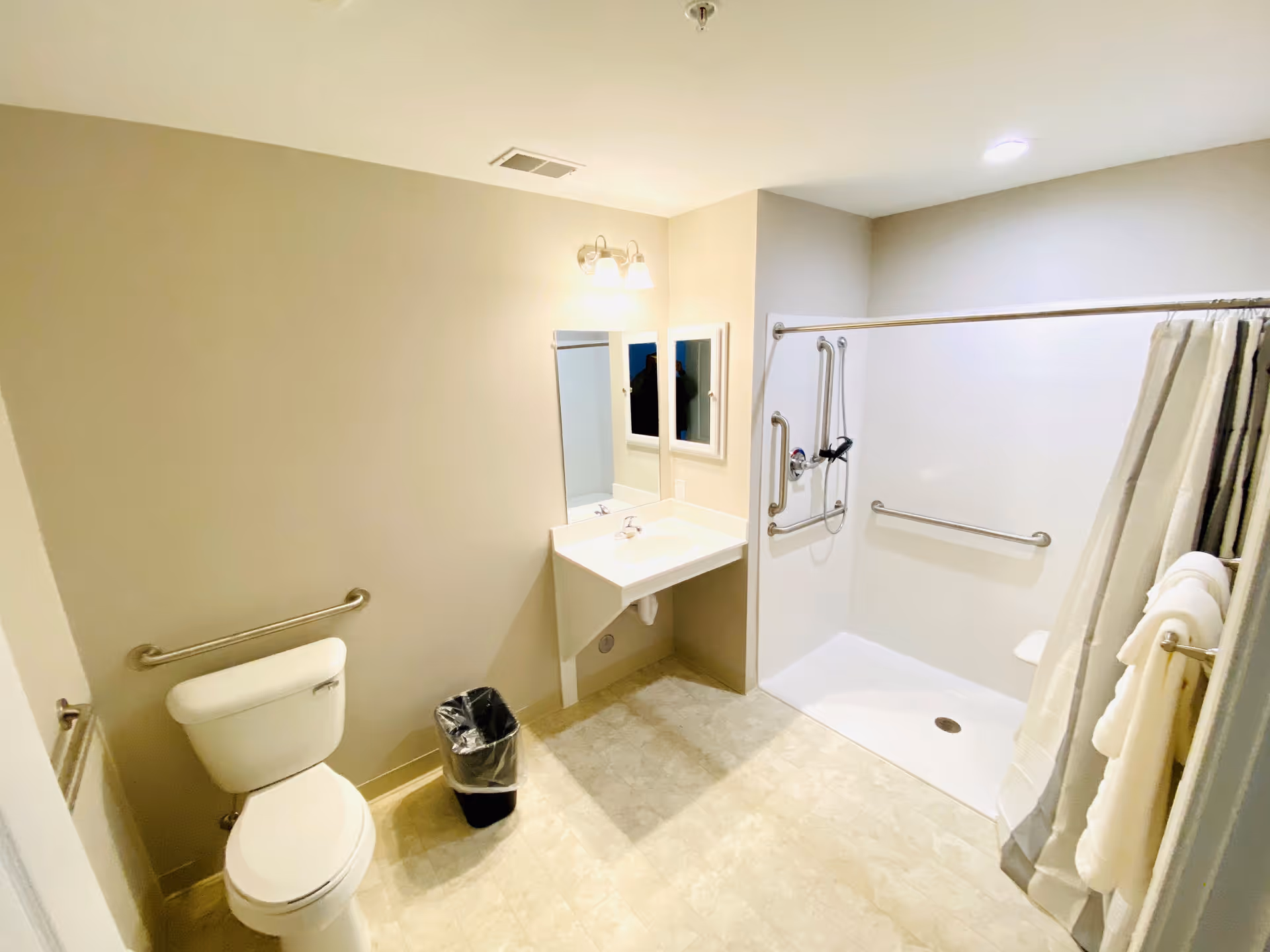 Bright accessible bathroom featuring a toilet, wall-mounted sink and mirror, and a roll-in shower with grab bars and a shower curtain.