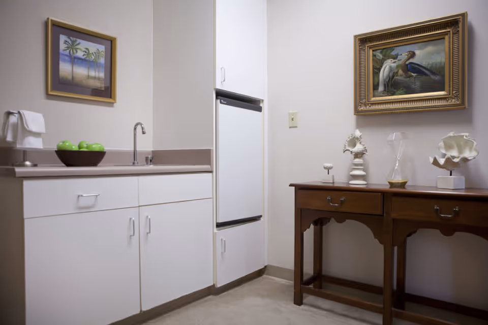 A small kitchenette area with white cabinets, a countertop with a sink, and a bowl of green apples. Next to it is a wooden table with decorative items including seashells and an hourglass. Two framed paintings hang on the walls, one depicting palm trees and the other showing birds near water.
