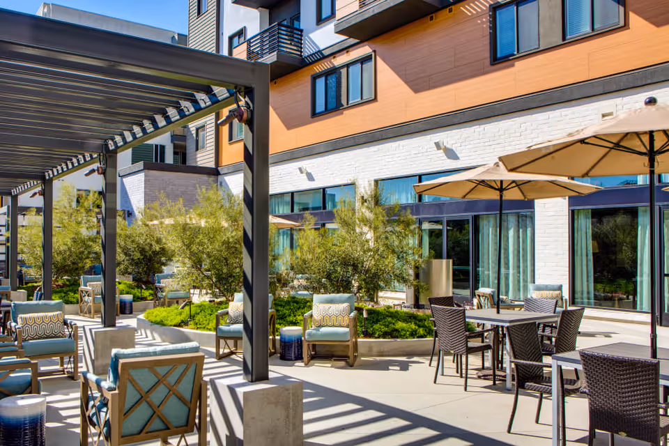Outdoor patio area at Sage Glendale featuring cushioned chairs, tables with umbrellas, greenery, and a modern building facade in the background under a clear sky.