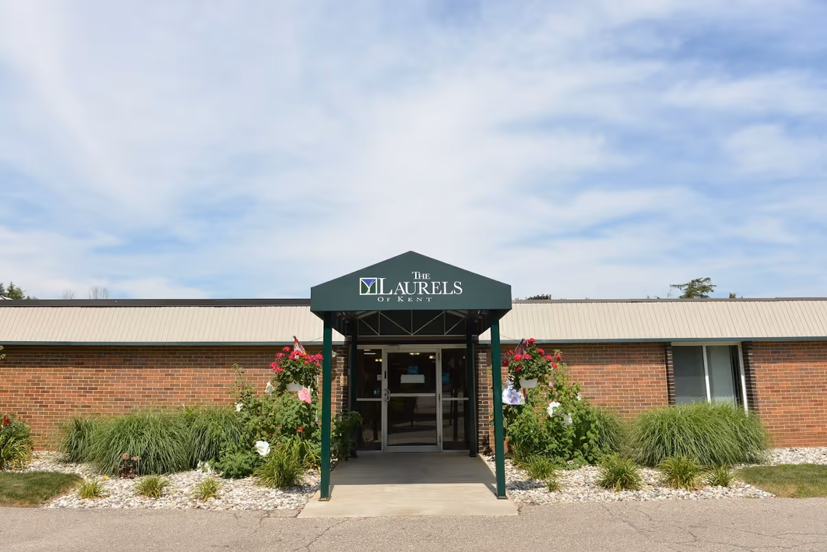 Front exterior view of a single-story brick building with a green awning that reads 'The Laurels of Kent'. There are plants and flowers on either side of the entrance, and the sky is partly cloudy.