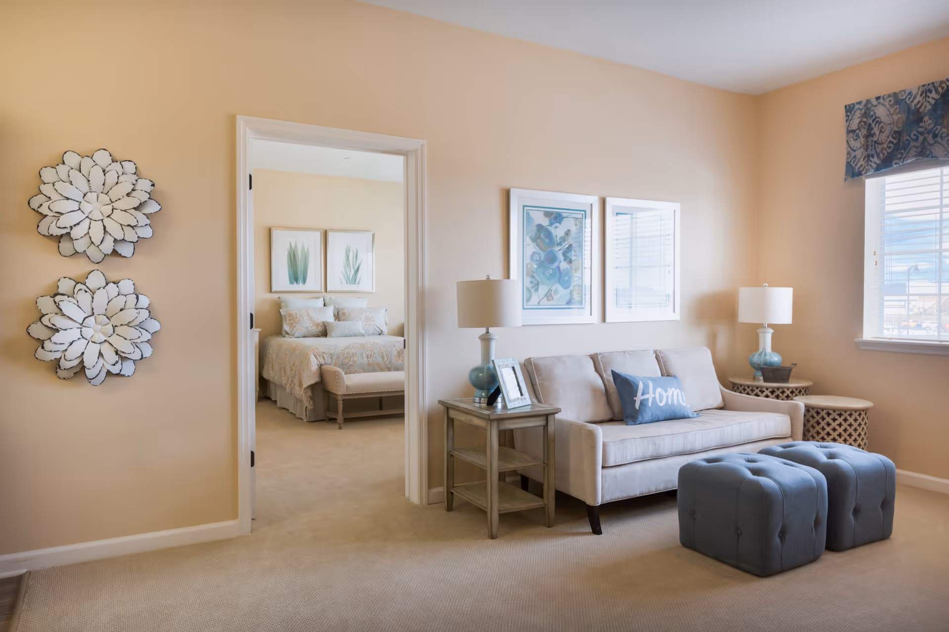 Sunlit living room with a beige sofa, two tufted blue ottomans, side tables and a view into a bedroom.