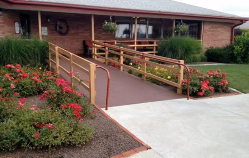 Exterior view of a single-story brick building with a wheelchair accessible ramp leading to the entrance. The ramp is flanked by wooden railings and surrounded by well-maintained flower beds with red and yellow flowers and green shrubs. The building has large windows and a covered porch area with hanging flower baskets.