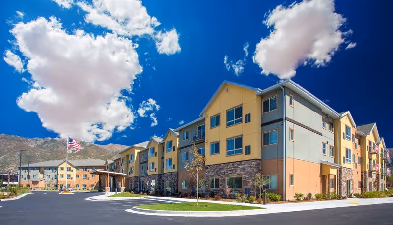 Exterior view of a multi-story senior living facility building under a bright blue sky with scattered clouds. The building features a combination of yellow, beige, and stone facade with multiple windows and a covered entrance. An American flag is visible near the entrance, and the surrounding area includes a paved driveway and some landscaping with small trees and shrubs.