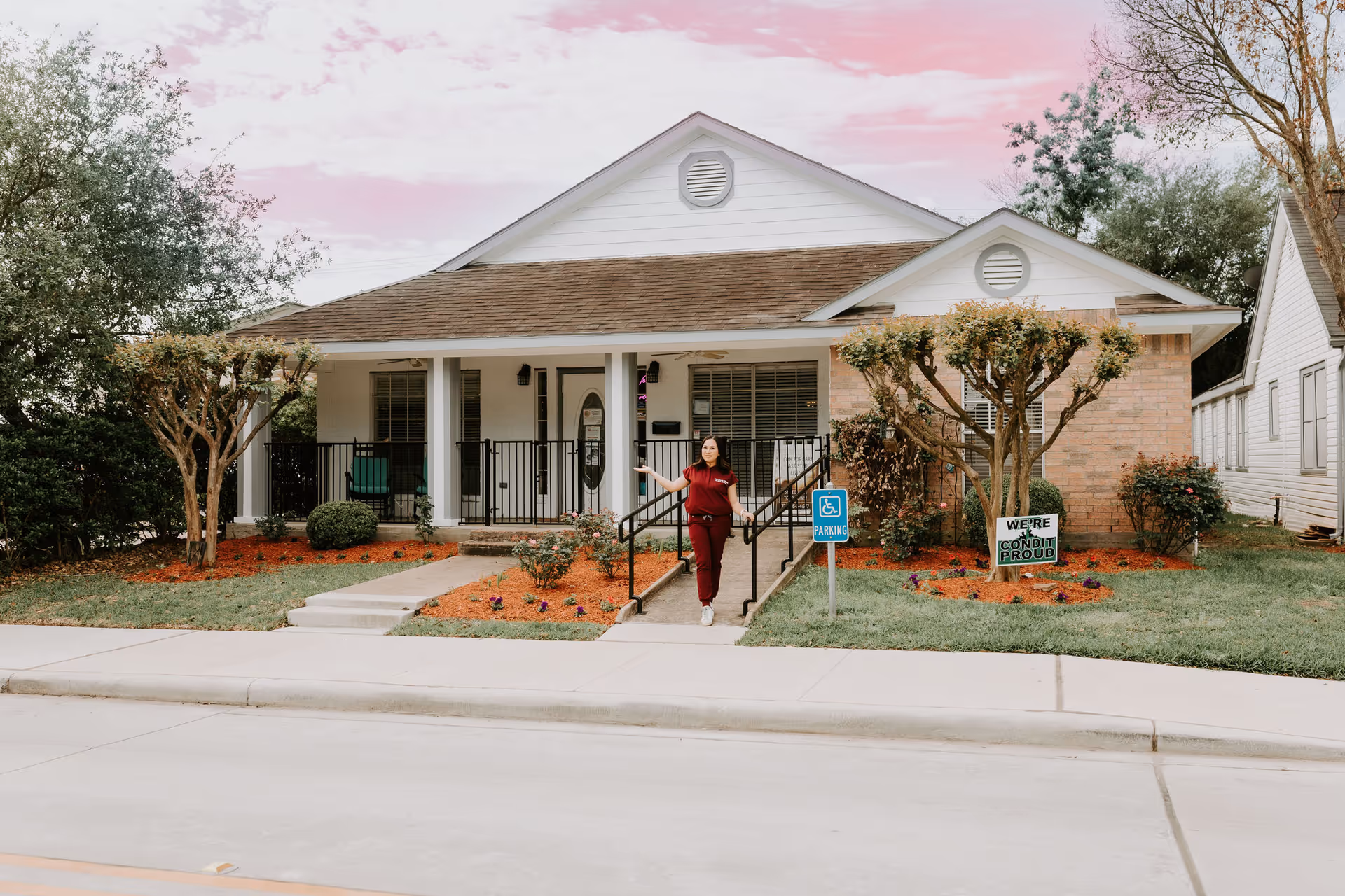 A person stands at the ramp of a one story senior facility with landscaped front yard.