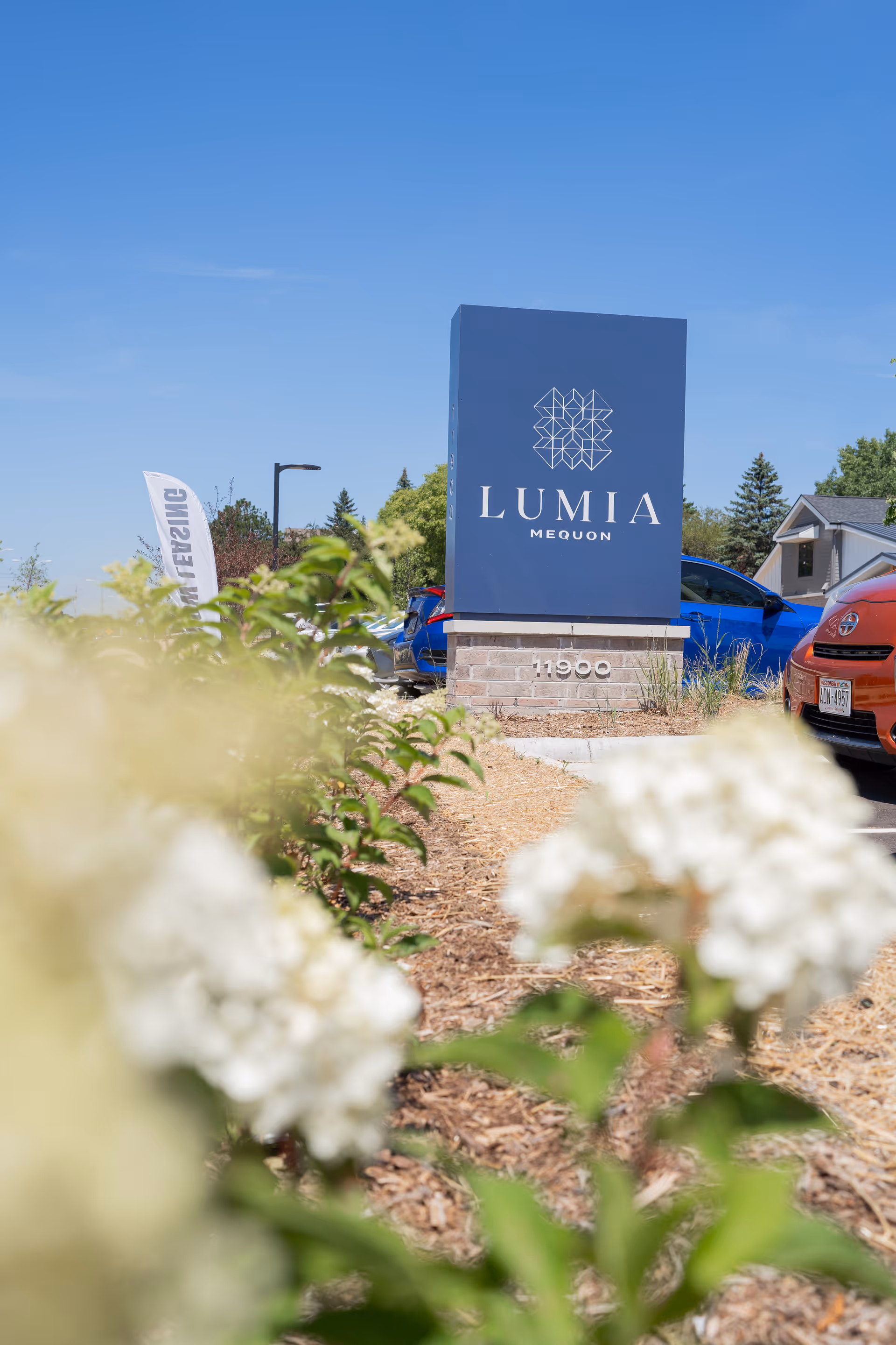 Outdoor view of the Lumia Mequon Assisted Living & Memory Care facility sign with plants and flowers in the foreground, parked cars, and a clear blue sky.