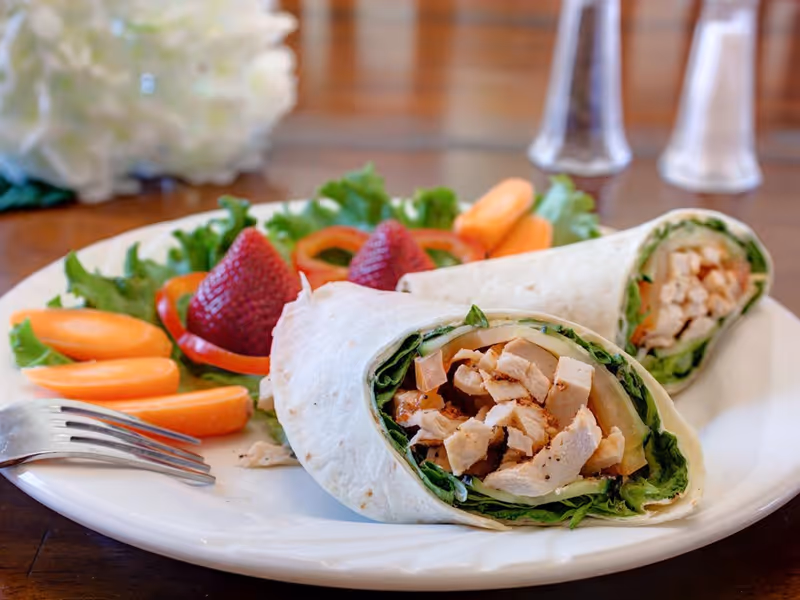 A plate with two chicken wraps filled with lettuce and other vegetables, accompanied by baby carrots, strawberries, and lettuce garnish. A fork is placed on the plate, and salt and pepper shakers are visible in the background on a wooden table.