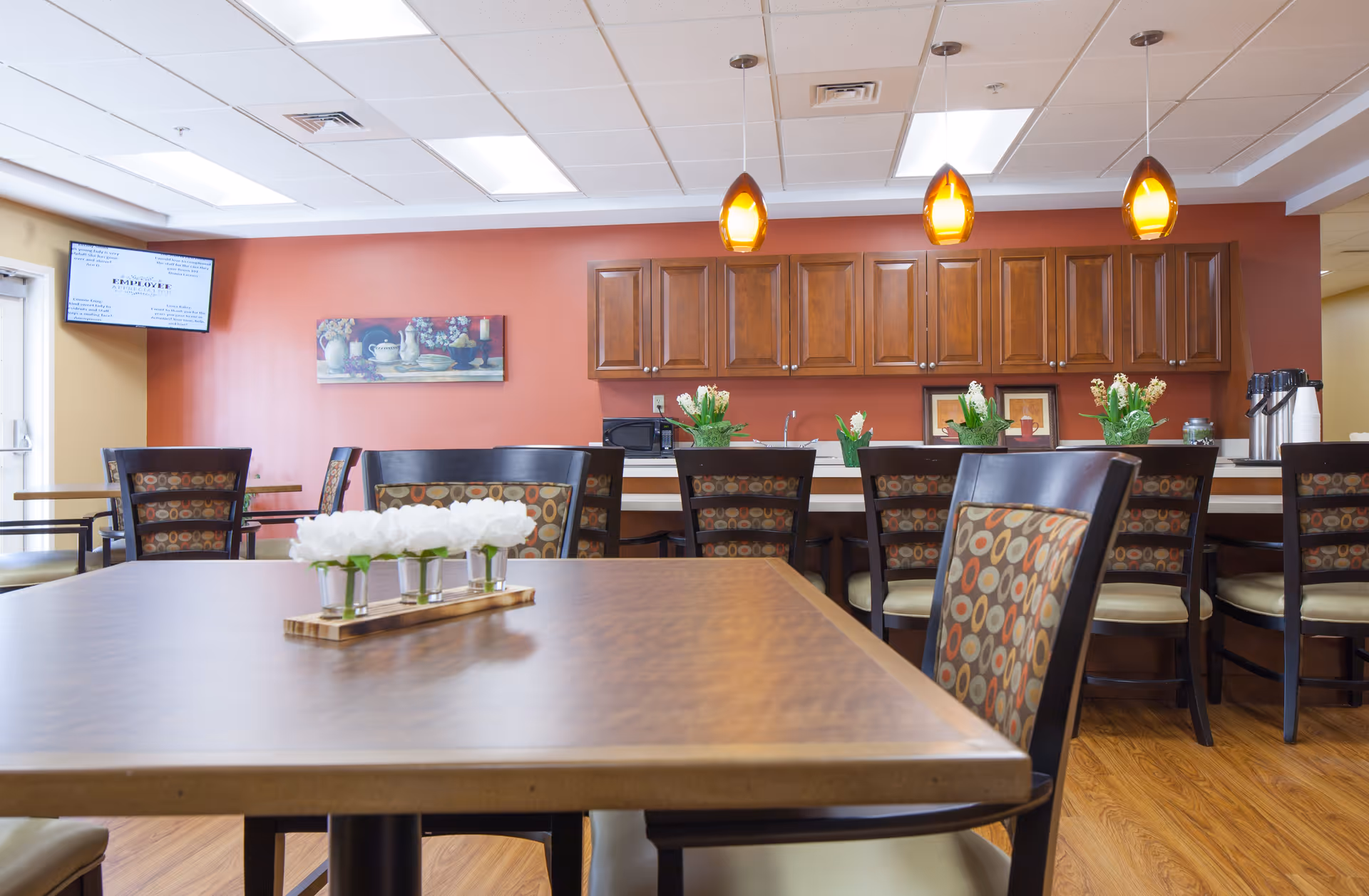 A dining area with wooden tables and chairs featuring patterned upholstery. The background shows a red wall with wooden cabinets, a microwave, and decorative plants on the counter. Three pendant lights hang from the ceiling, and a TV screen is mounted on the left wall.