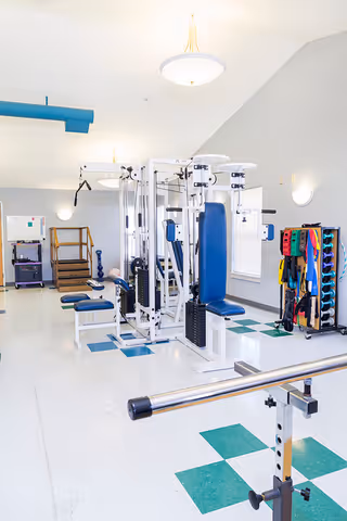 Bright physical therapy/fitness room with multi-station weight machines, parallel bars, and a rack of colorful exercise bands.