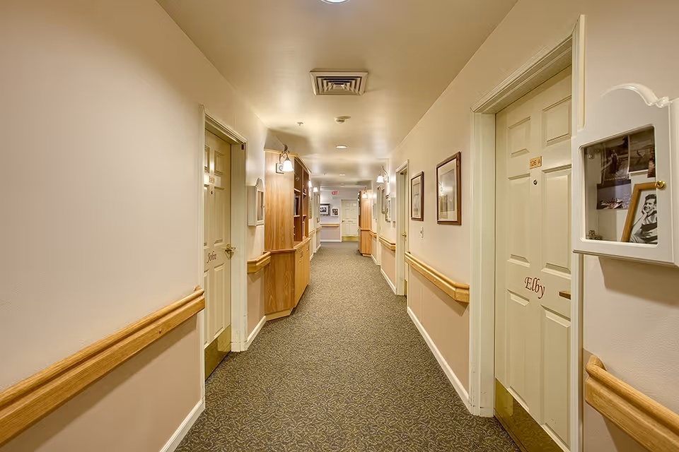 A well-lit hallway in a senior living facility with beige walls, carpeted floor, wooden handrails, and several closed doors labeled with names. There are framed pictures on the walls and built-in wooden cabinets along the corridor.